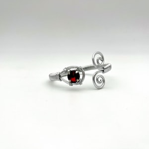 Könnte beinhalten: Ein silberfarbenes Drahtarmband mit einem zentralen, runden, tiefroten Edelstein. Das Armband hat ein Spiraldesign an einem Ende und eine käfigartige Fassung für den Edelstein. Das Design ist minimalistisch und elegant.