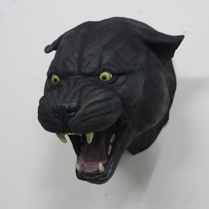 Puede incluir: Escultura de cabeza de pantera negra con la boca abierta y dientes visibles. La pantera tiene ojos verdes brillantes y está montada en una pared blanca. La escultura es una representación detallada de la cara de una pantera.