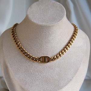 Puede incluir: Un collar de cadena dorada con un diseño de eslabones grandes. El collar presenta un colgante ovalado central con las letras "CD" en tono dorado a juego. El collar se exhibe sobre un busto de color crema.