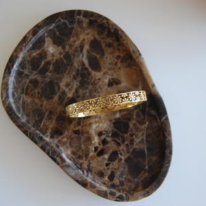 Könnte beinhalten: Ein goldfarbenes Armband mit floralem Muster liegt auf einem braunen und beigefarbenen Marmortablett. Das Armband ist ein durchgehendes Band mit einem sich wiederholenden Design aus durchbrochenen Blumen. Das Tablett hat eine glatte, polierte Oberfläche.