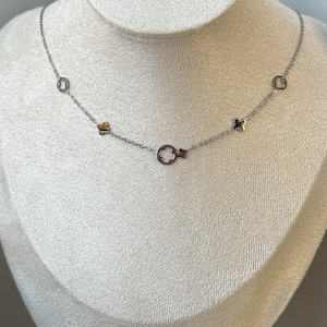 Op de afbeelding: Een zilverkleurige ketting met meerdere klaver- en cirkelvormige bedels. De ketting wordt getoond op een witte mannequin. De bedels hebben gouden en zilveren accenten. De ketting is delicaat en elegant.