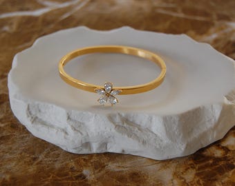 Op de afbeelding: Een goudkleurige armband met een bloemvormige cluster van heldere edelstenen. De armband ligt op een witte, onregelmatig gevormde steen. De achtergrond is een gevlekte bruin-beige oppervlak.