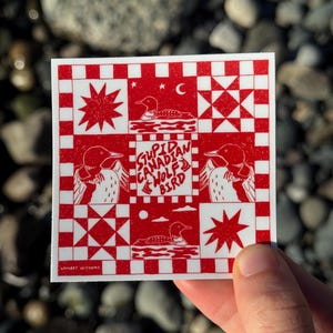 May include: Square sticker with a red and white pattern featuring loon illustrations, stars, and a crescent moon. The text "STUPID CANADIAN WOLF BIRD" is in the center. The sticker is held by a hand.