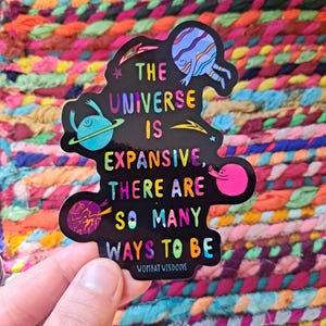 May include: A black sticker with a colorful design featuring planets and stars. The text on the sticker reads "The Universe is Expansive, There are So Many Ways to Be." The sticker also includes the text "Wombat Wisdoms".