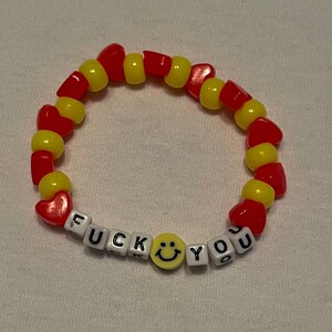 May include: A red and yellow beaded bracelet with the words "F U C K YOU" spelled out in white beads and a yellow smiley face bead.