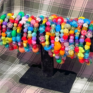 Può includere: Una collezione di braccialetti colorati con perline. I braccialetti presentano una varietà di forme, tra cui fiori, cuori e stelle, in tonalità di rosso, arancione, giallo, verde, blu e viola. I braccialetti sono esposti su un supporto nero.