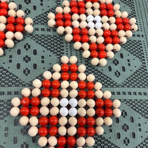 May include: Close-up of several diamond-shaped trivets. Each trivet is made of small, round beads in red, white, and natural wood colors. The trivets are arranged on a green, patterned surface.