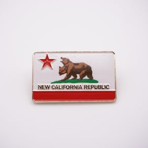 May include: A gold-colored pin with a red star, a brown bear, and green grass on a white background. The text "NEW CALIFORNIA REPUBLIC" is printed in black below the bear.
