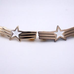 May include: Two gold-toned hair clips, each featuring a white star and a stylized comet tail design. The star is centered, with the tail composed of three parallel lines. The clips are set against a white background.