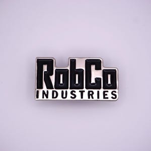 May include: A silver-colored pin with the text "RobCo INDUSTRIES" in black, block letters. The pin has a rectangular shape with a raised, glossy black text and a silver border. The pin is on a light gray background.
