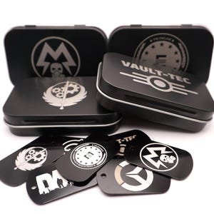 May include: A collection of black metal rectangular tins and matching dog tags featuring various white engraved logos, including Vault-Tec, The Enclave, and other symbols. The items are arranged in a group with the tins standing upright behind the tags.