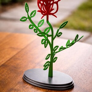 May include: A decorative metal rose sculpture. The red rose is atop a green stem with green leaves. The sculpture is on a black oval base. The background is blurred.
