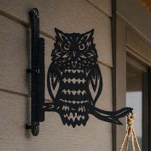 Op de afbeelding: Zwarte metalen uilvormige plantenhouder bevestigd aan een lichtgekleurde muur. De uil zit op een tak, met een hangende plantenbak gevuld met groene planten eronder. De plantenbak heeft een rustieke bruine kleur.