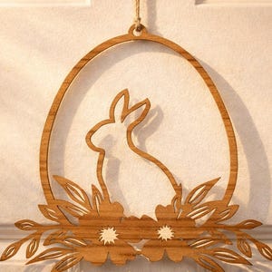 Op de afbeelding: Houten paas-ei-vormige wandhanger met een konijnensilhouet aan de binnenkant. De bruine decoratie heeft bloemen en bladeren uitsparingen aan de onderkant en wordt opgehangen door een natuurlijke touw. Een decoratief stuk voor het voorjaarsseizoen.