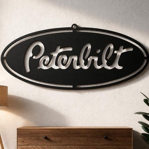 Peut inclure: Panneau Peterbilt en métal noir de forme ovale avec le nom de la marque découpé en blanc. Le panneau est accroché à un mur blanc au-dessus d'une commode en bois. Une lampe de sol et une plante en pot sont également dans l'image.