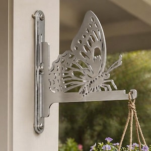Op de afbeelding: Een zilveren metalen plantenbak in de vorm van een vlinder is aan een witte muur bevestigd. Het vlinderontwerp is uitgesneden en een hangmand met paarse bloemen is eraan opgehangen. De hanger is ontworpen voor buitengebruik.