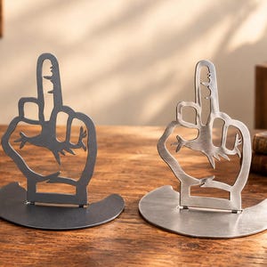 May include: Three metal bookends, each featuring a cutout design of a hand making a middle finger gesture. The set includes one black, one dark grey, and one silver-toned piece, all mounted on a semi-circular base for stability on a flat surface.