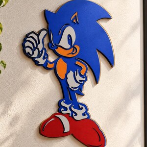Könnte beinhalten: Ein wandmontierter, mehrschichtiger Holzausschnitt der Figur Sonic the Hedgehog mit einem leuchtend blauen Körper, pfirsichfarbener Haut an Armen und Schnauze, weißen Handschuhen und großen roten Schuhen mit weißen Akzenten, in einer Pose mit erhobenem Daumen.