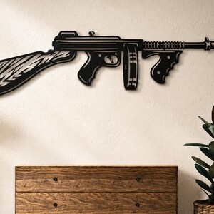 May include: A black metal wall decoration in the shape of a Thompson submachine gun. The artwork shows a detailed outline of the firearm, including the stock, barrel, and magazine. It is mounted on a neutral-coloured wall.
