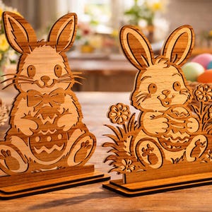 May include: Two wooden Easter bunny decorations. One bunny has a bow tie and holds an Easter egg. The other bunny is surrounded by flowers and grass and also holds an egg. Both are on a wooden base.