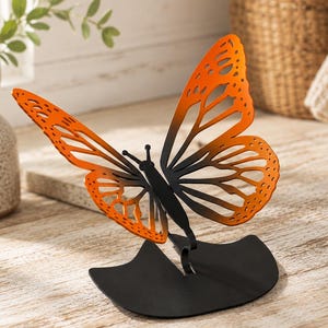 May include: A metal butterfly sculpture featuring orange and black wings with a cut-out pattern. The butterfly is attached to a solid black, curved base that allows it to stand upright. The wings transition from a vibrant orange to black.