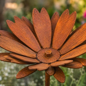 May include: A large, rusty metal flower sculpture with layered petals and a central bolt. The flower is a warm, earthy brown color and is set against a blurred background of green foliage and colorful flowers.