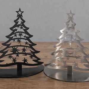 Puede incluir: Dos adornos de árbol de Navidad de metal. Uno es negro, el otro plateado. Ambos tienen una estrella en la parte superior y recortes de estrellas en todo el diseño del árbol. Se apoyan sobre una base redonda y se exhiben sobre una superficie de madera.