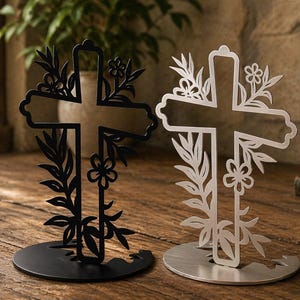 May include: Two decorative crosses, one black and one silver, are displayed on a wooden surface. Each cross is adorned with floral and leaf cutouts. The crosses stand on circular bases, with a natural background.