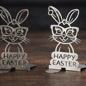 Puede incluir: Dos figuras de conejos de Pascua de metal con gafas y las palabras "HAPPY EASTER" recortadas. Los conejos tienen orejas largas y están de pie sobre una pequeña base. Las figuras son plateadas y se asientan sobre una superficie marrón oscuro.