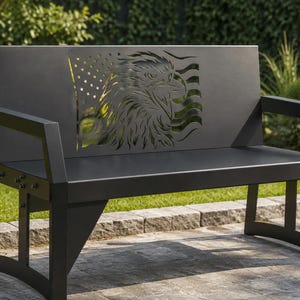 Puede incluir: Un banco de metal negro con respaldo con un diseño recortado de un águila y la bandera estadounidense. El banco tiene reposabrazos y está colocado en un patio de piedra con un fondo de hierba.