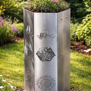 May include: A tall, brushed metal planter with laser-cut floral and bee designs. The hexagonal planter is filled with soil and topped with pink flowers. The planter is set on a grassy lawn.