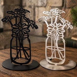 May include: Two metal daisy flower sculptures in mason jar shapes. One is black, the other is silver. Both are on round bases. The background includes a brick wall and a wooden table.