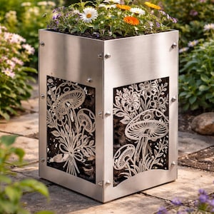 Könnte beinhalten: Ein quadratischer Pflanzkasten aus Edelstahl, gefüllt mit Erde und bunten Blumen. Die Seiten weisen dekorative Ausschnitte von Pilzen und Blättern auf. Der Pflanzkasten befindet sich im Freien, umgeben von Grün und Steinplatten.