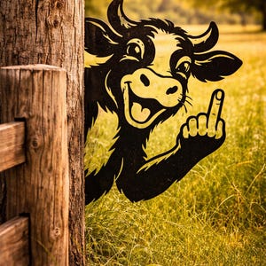 May include: A black and yellow cow cutout giving the middle finger, peeking from behind a tree and wooden fence. The cow is smiling with a yellow face and horns. The background is a field of tall grass.