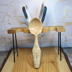 May include: A handcrafted wooden spoon with an ornate, engraved design on the handle. The spoon is displayed on a small wooden table with black metal legs, and two decorative feathers are positioned behind it. The background features a marble-like pattern.