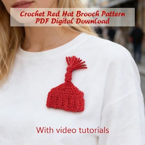 May include: A red crochet hat brooch pinned to a white shirt. The brooch has a textured, knitted appearance with a tassel detail. Text on the image reads "Crochet Red Hat Brooch Pattern PDF Digital Download" and "With video tutorials."