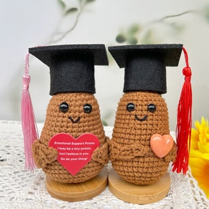 May include: Two brown crocheted potato figures wearing graduation caps and holding hearts. One cap has a pink tassel and the other has a red tassel. The potato on the left is holding a red heart with the text "Emotional Support Potato I may be a tiny potato, but I believe in you. Go do your thing!"