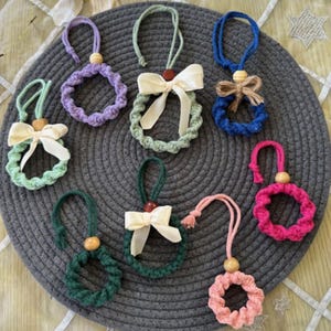 May include: Eight handmade macrame wreaths in various colours, including purple, green, blue, pink, and red. Each wreath has a wooden bead and a ribbon bow. The wreaths are displayed on a grey circular mat.