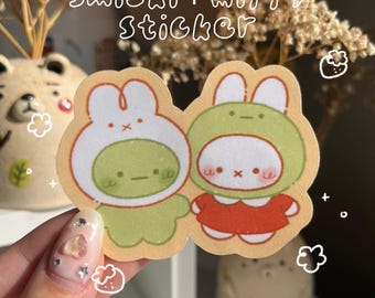 May include: A sticker featuring two cartoon characters, one in a green frog costume and the other in a white bunny costume, both with smiling faces. The sticker has a light tan border and the text "smiski + miffy sticker" in white script.