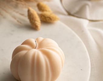 May include: A pale peach-colored pumpkin-shaped candle with a single wick on a white marble surface.