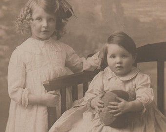 Könnte beinhalten: Ein Vintage-Foto in Sepia zeigt zwei junge Mädchen. Eines mit lockigem Haar und einem weißen Kleid steht neben einem sitzenden Mädchen in einem ähnlichen Kleid. Das sitzende Mädchen hält einen strukturierten Gegenstand. Sie sitzen auf einer Holzbank.