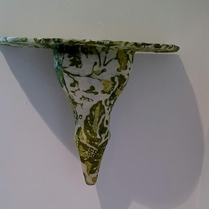 May include: A wall-mounted shelf featuring a semi-circular top surface and a tapered, cone-shaped base. The entire structure is covered in a white paper-like material decorated with a repeating green botanical pattern of leaves and small dots.