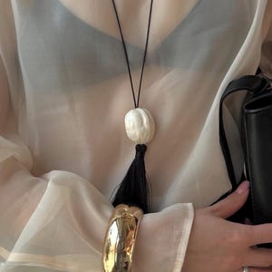 Könnte beinhalten: Eine lange Halskette mit einer dünnen schwarzen Kordel, einer glatten weißen ovalen Perle und einer schwarzen Quaste. Die Kette wird über einem transparenten beigen Langarmshirt mit schwarzer Unterwäsche getragen, ergänzt durch einen goldenen Armreif und eine schwarze Handtasche.