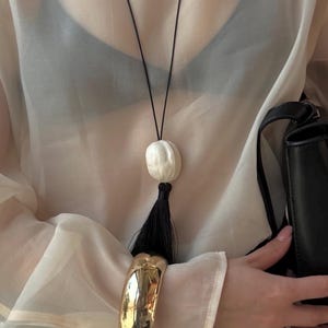 May include: A long necklace featuring a thin black cord, a smooth white oval bead, and a black tassel. The necklace is worn over a sheer beige long-sleeve top with a black bra underneath. A wide, polished gold-tone metal cuff bracelet is on the wrist.