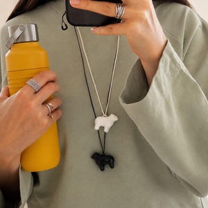 May include: A sage green long-sleeve shirt, a yellow metal water bottle with a silver cap, and two necklaces featuring small sheep pendants, one white and one black, on thin cords. The wearer has silver rings on their fingers and holds a black phone.