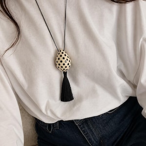 May include: A long black necklace with a cream-colored, teardrop-shaped pendant adorned with black polka dots. The pendant is paired with a black tassel. The necklace is worn over a white long-sleeved shirt and blue jeans.