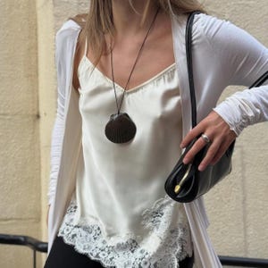 May include: A black shell pendant necklace hangs over a white camisole with lace trim. A black purse with a gold zipper is held in the hand. A white cardigan is worn over the camisole.