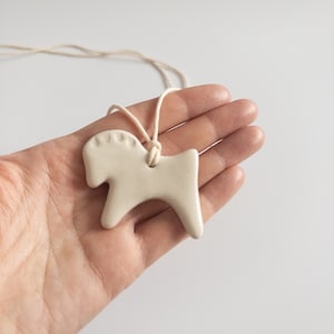 Op de afbeelding: Een ketting met een witte keramische paardenhanger met een eenvoudig ontwerp. De hanger is bevestigd aan een wit touwtje.