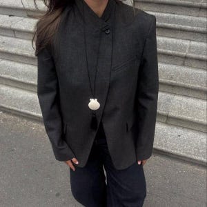 Könnte beinhalten: Ein dunkelgrauer Blazer mit einem einzigartigen Kragen und einem Ein-Knopf-Verschluss. Ein weißer Muschel-Anhänger hängt an einer schwarzen Kordel. Der Blazer hat eine Tasche auf der linken Seite und wird mit dunkelblauen Jeans getragen.