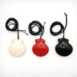 Könnte beinhalten: Drei muschelförmige Anhänger an schwarzen Kordelketten. Die Anhänger bestehen aus einem glatten, glänzenden Material in Weiß, Rot und Schwarz. Jedes Stück zeigt ein klassisches Fächerdesign einer Jakobsmuschel mit vertikalen Rillen.